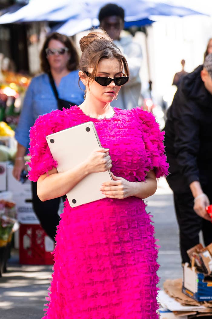 Selena Gomez in a textured dress, holding a laptop under her arm, wearing sunglasses while walking outdoors