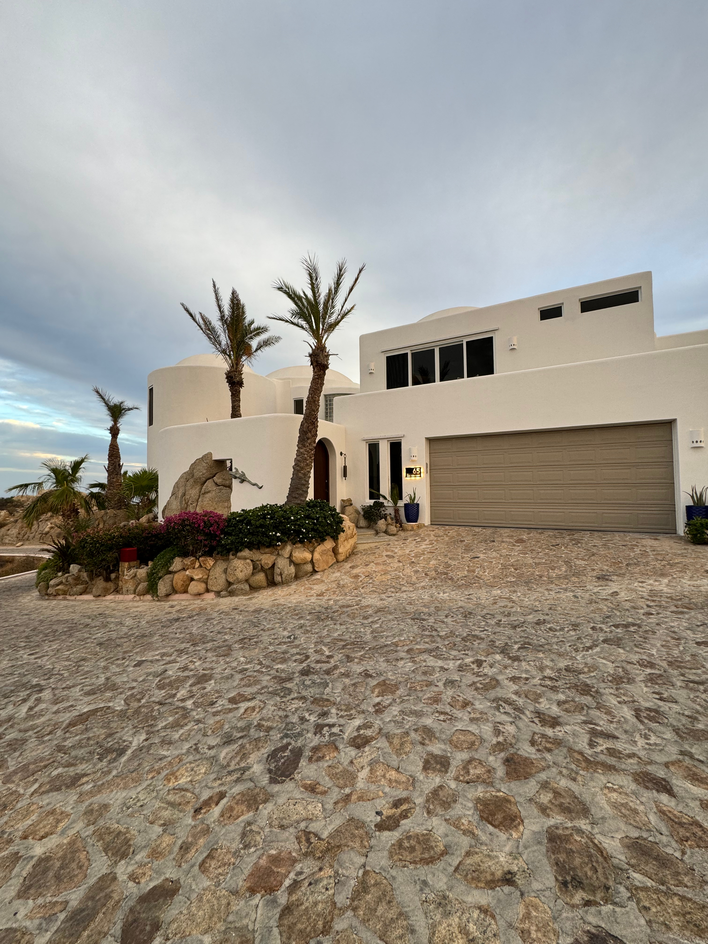 Exterior of a white, modern Mediterranean-style house with palm trees and a cobblestone driveway