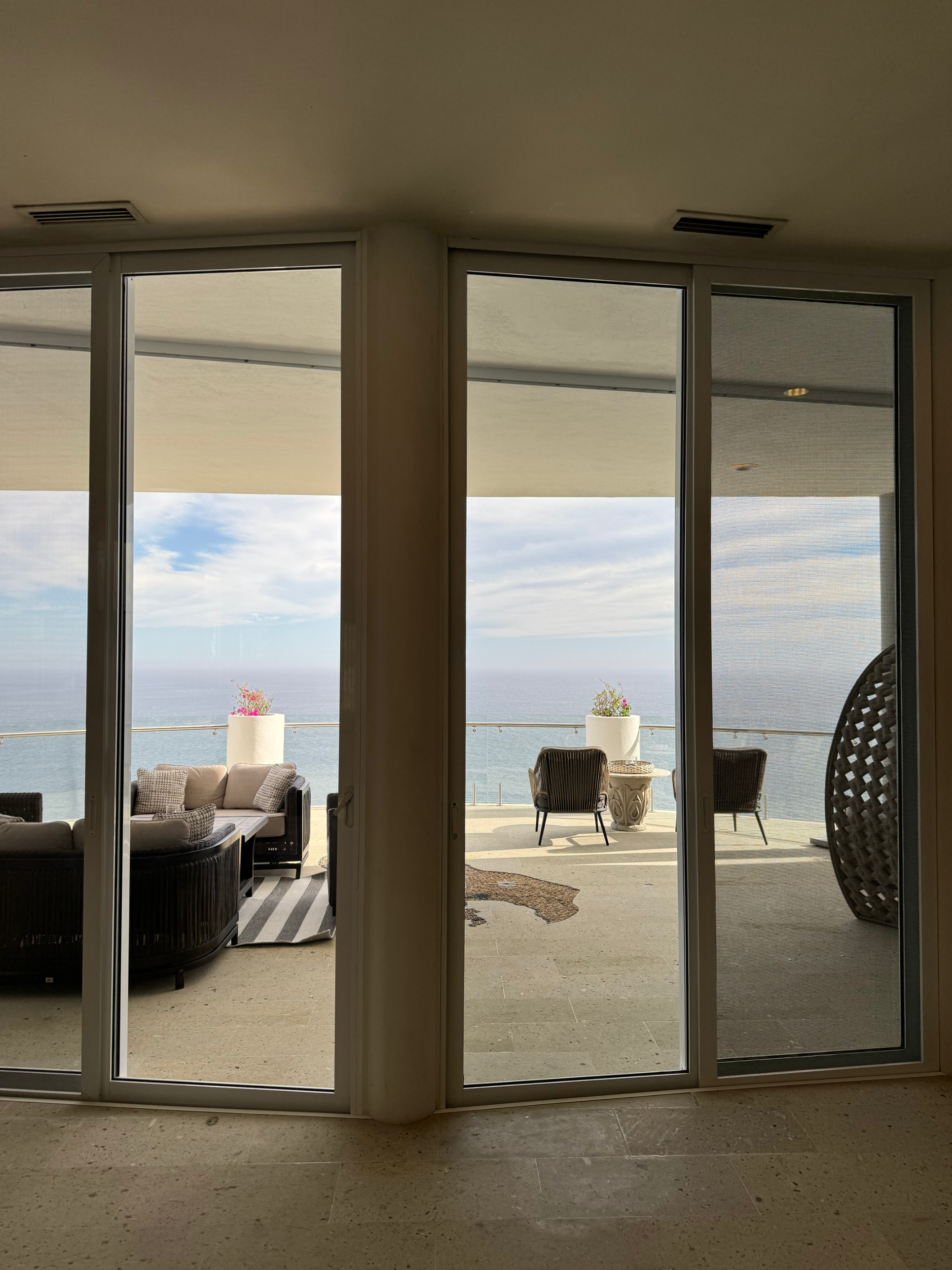 View through large glass doors to a modern balcony with outdoor furniture, overlooking the ocean under a partly cloudy sky