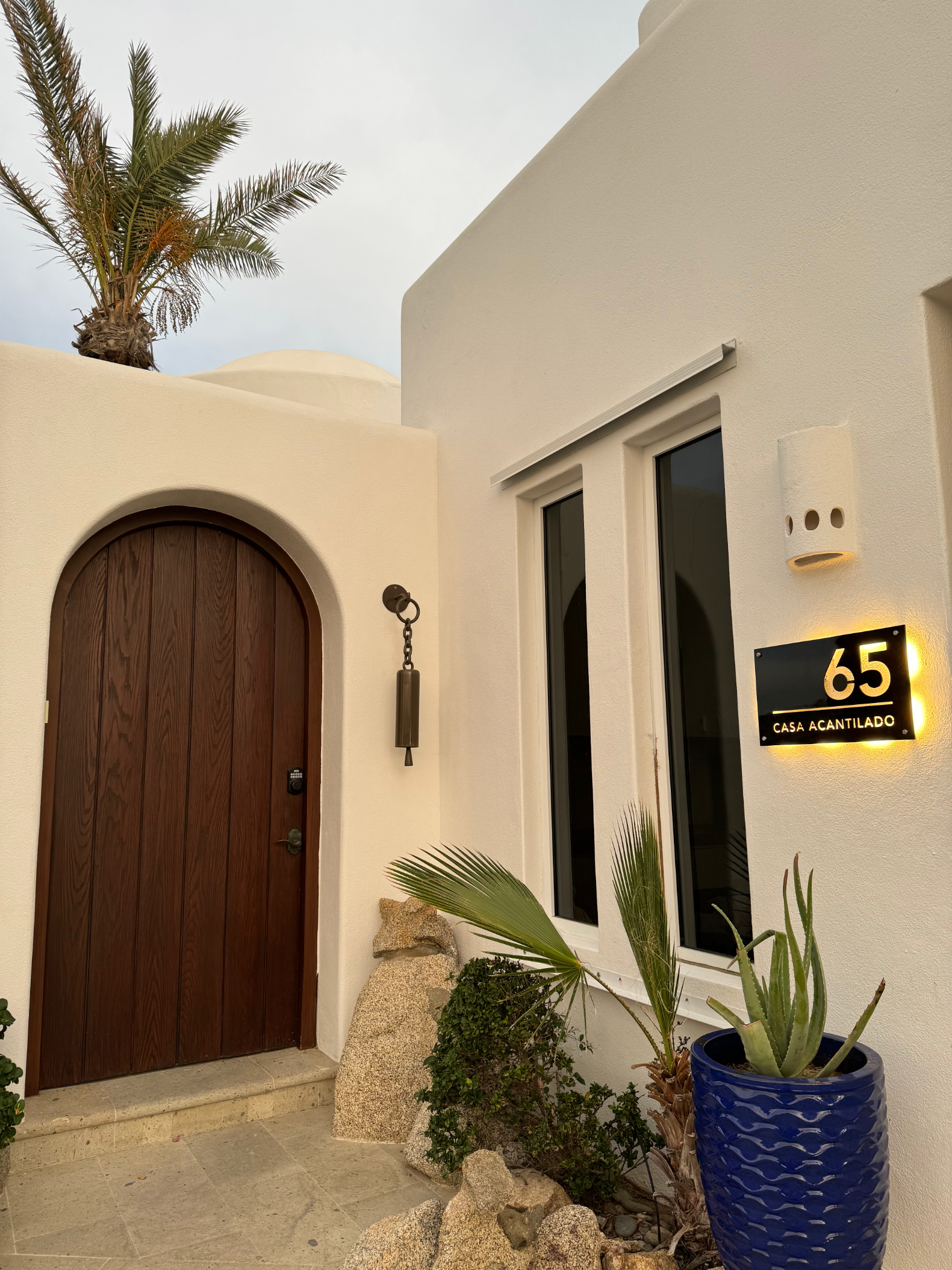 Exterior of a Mediterranean-style villa with a wooden arched door, plants, and a sign reading "65 CASA ACANTILADO," located in a travel destination