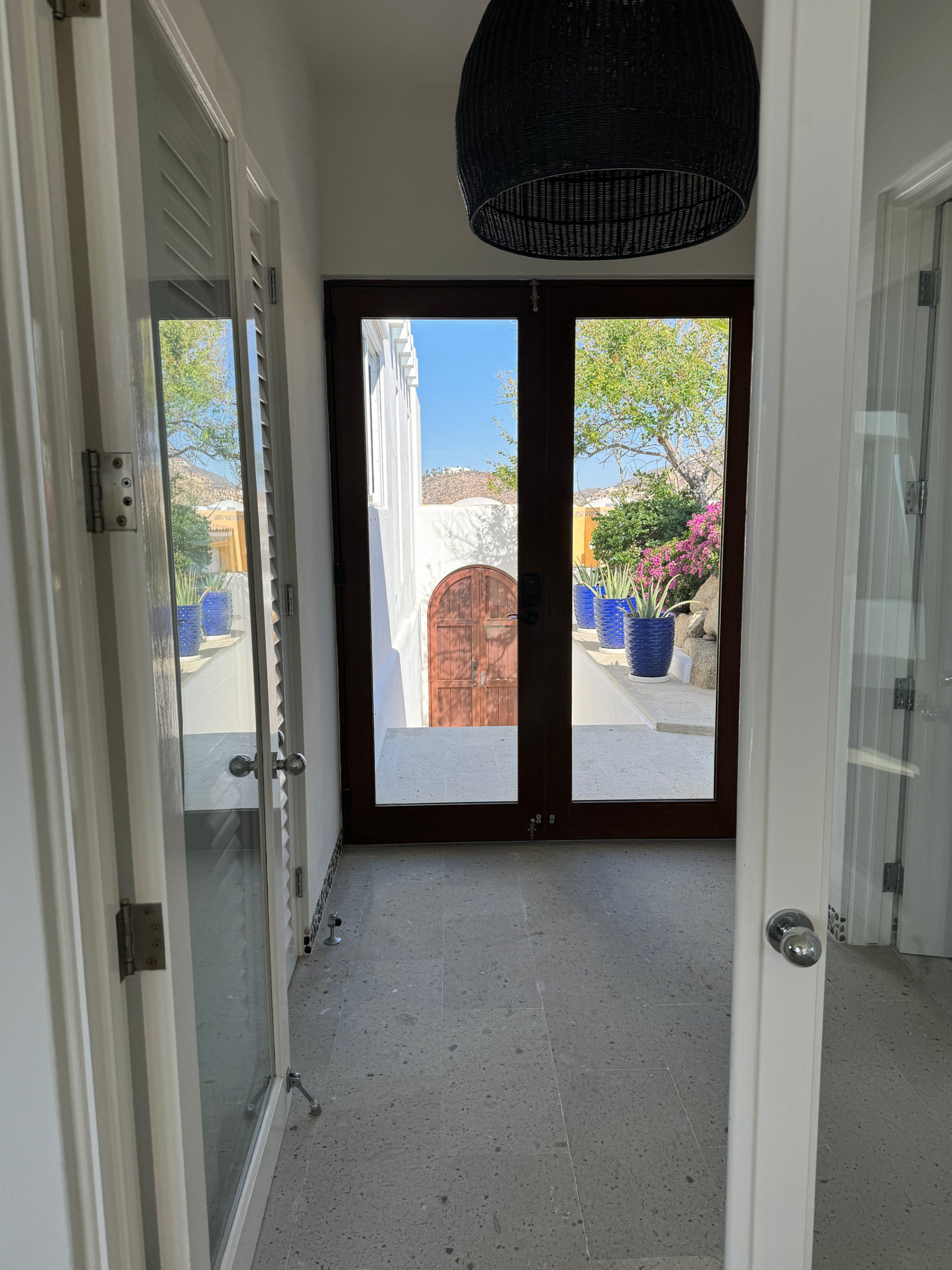 View through glass doors to a sunlit, vibrant patio with potted plants and a gate. The image showcases a serene, inviting outdoor space