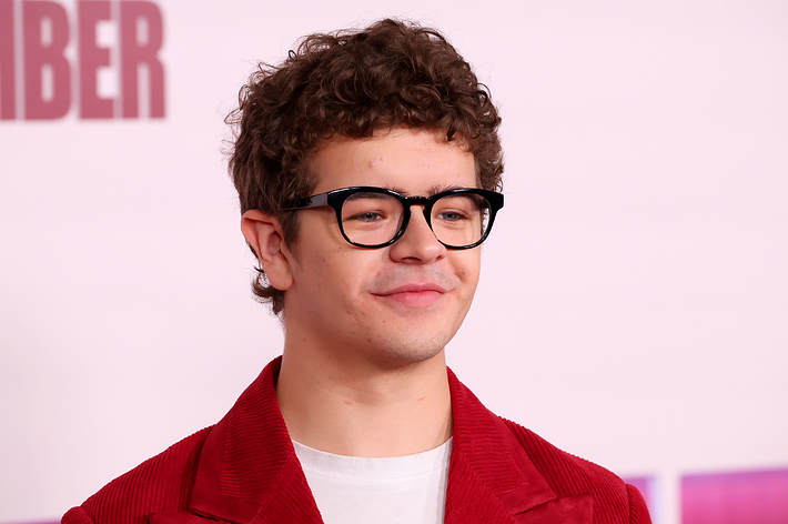 Gaten Matarazzo at an event, wearing black-frame glasses, a white shirt, and a casual red jacket