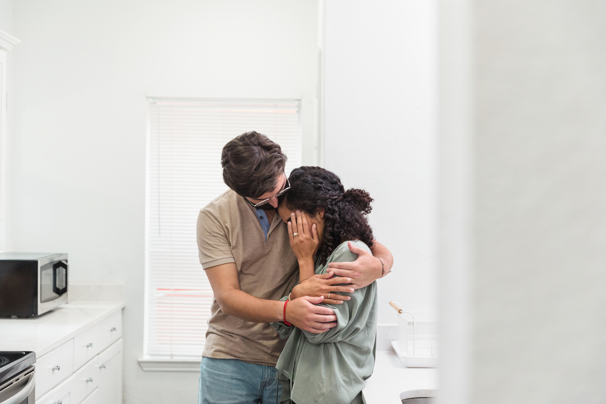 A man and a woman embrace in a kitchen, with the woman’s face partially covered by her hands. The setting suggests comfort and intimacy