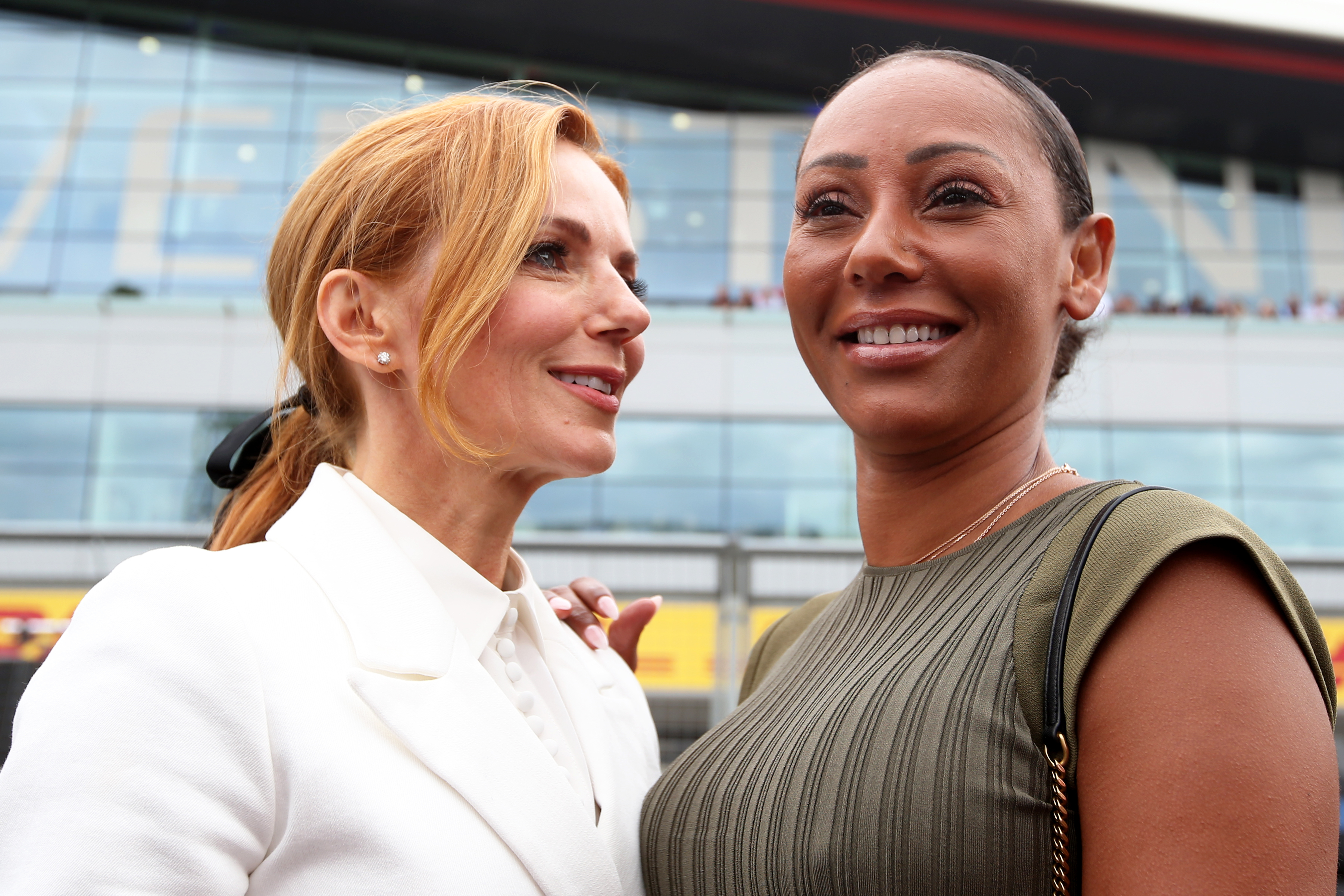 Geri Halliwell in a white outfit and Mel B in a green dress smiling together, standing outside a modern glass building