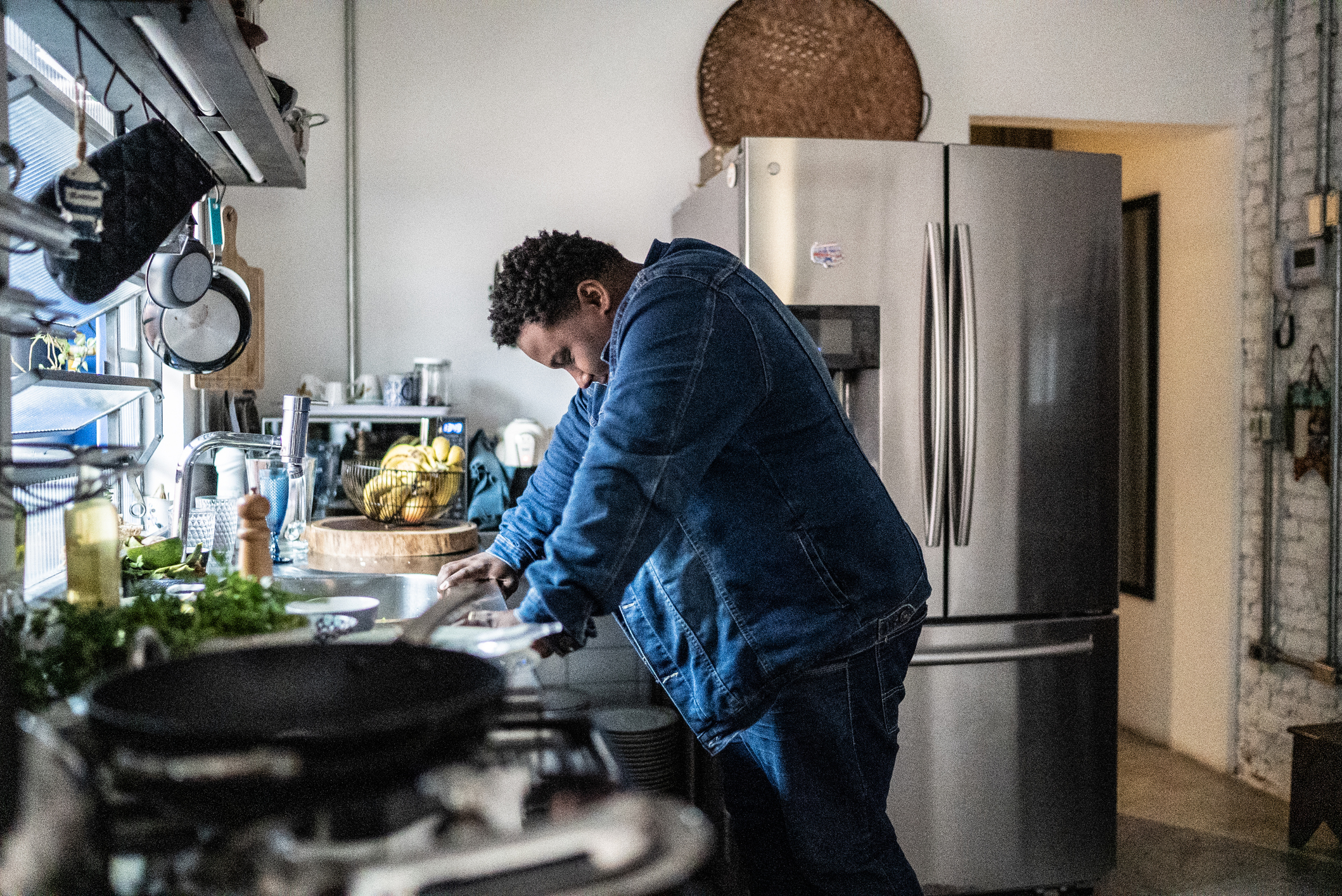 Man in denim jacket leans over a kitchen counter, focusing on something out of frame. Stainless steel refrigerator in the background