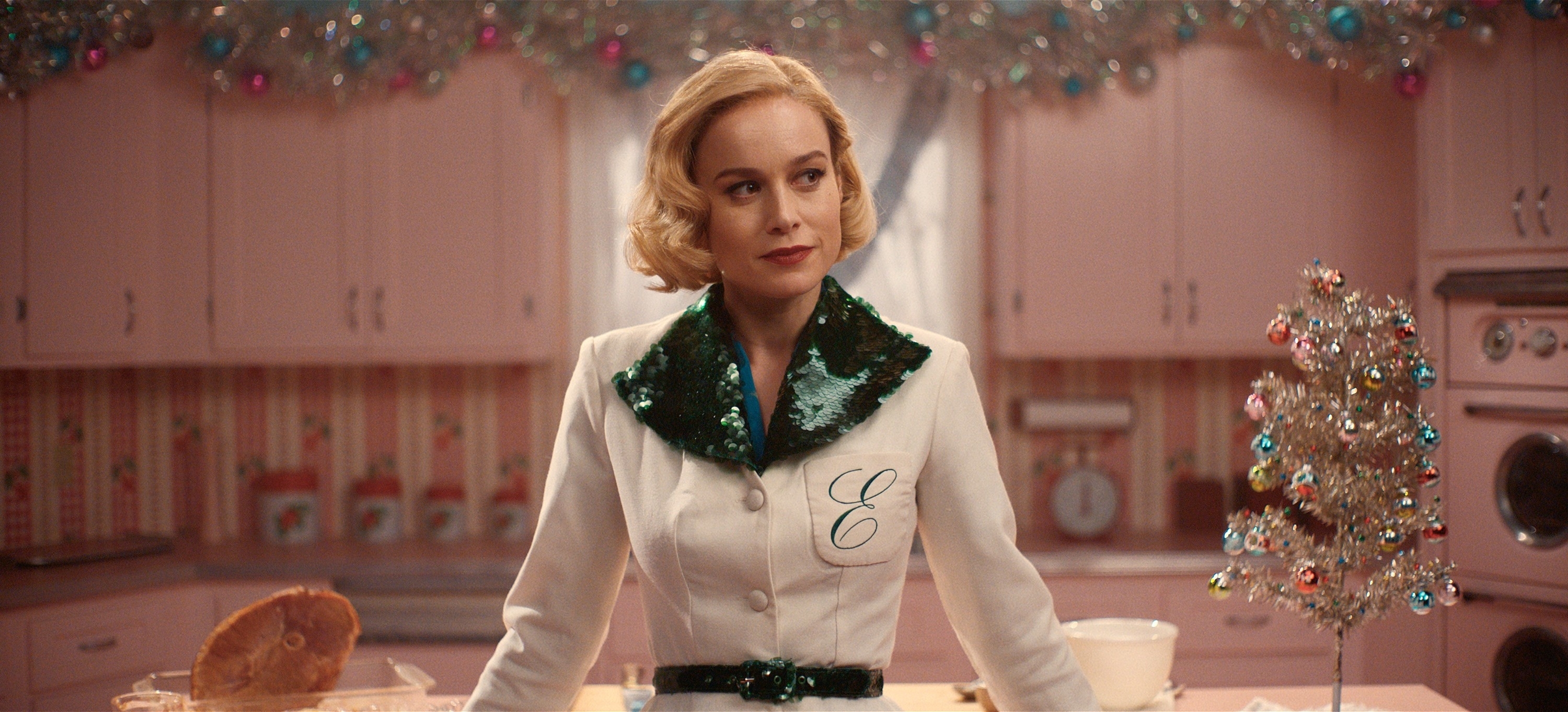 Brie Larson stands in a retro-style kitchen, wearing a white coat with green sequined detailing and an "E" monogram. A small decorated silver Christmas tree is on the counter