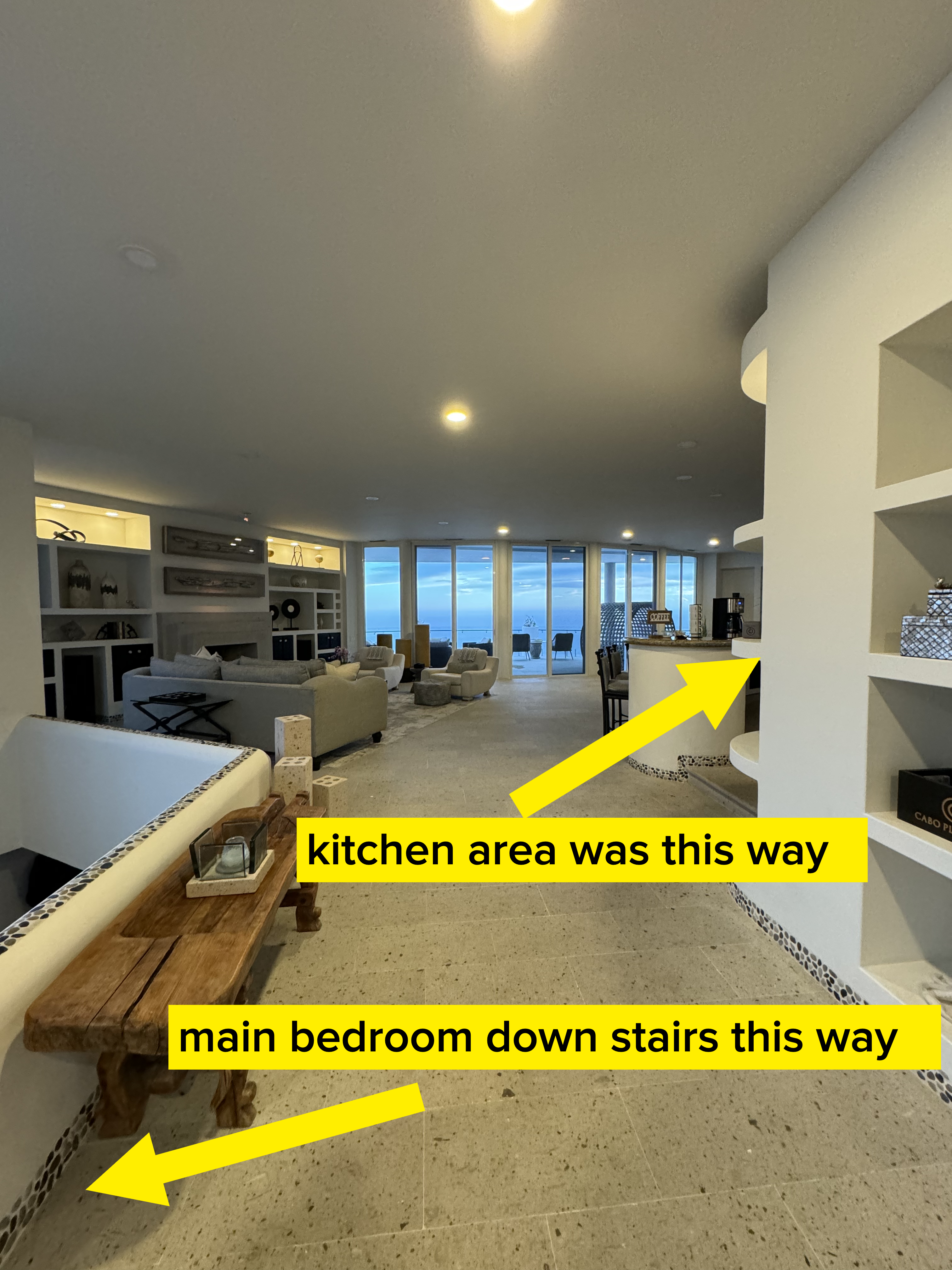 Spacious living room with modern decor, white furniture, and a view of the ocean through large windows. Shelves are built into the walls, and a wooden bench is in the foreground