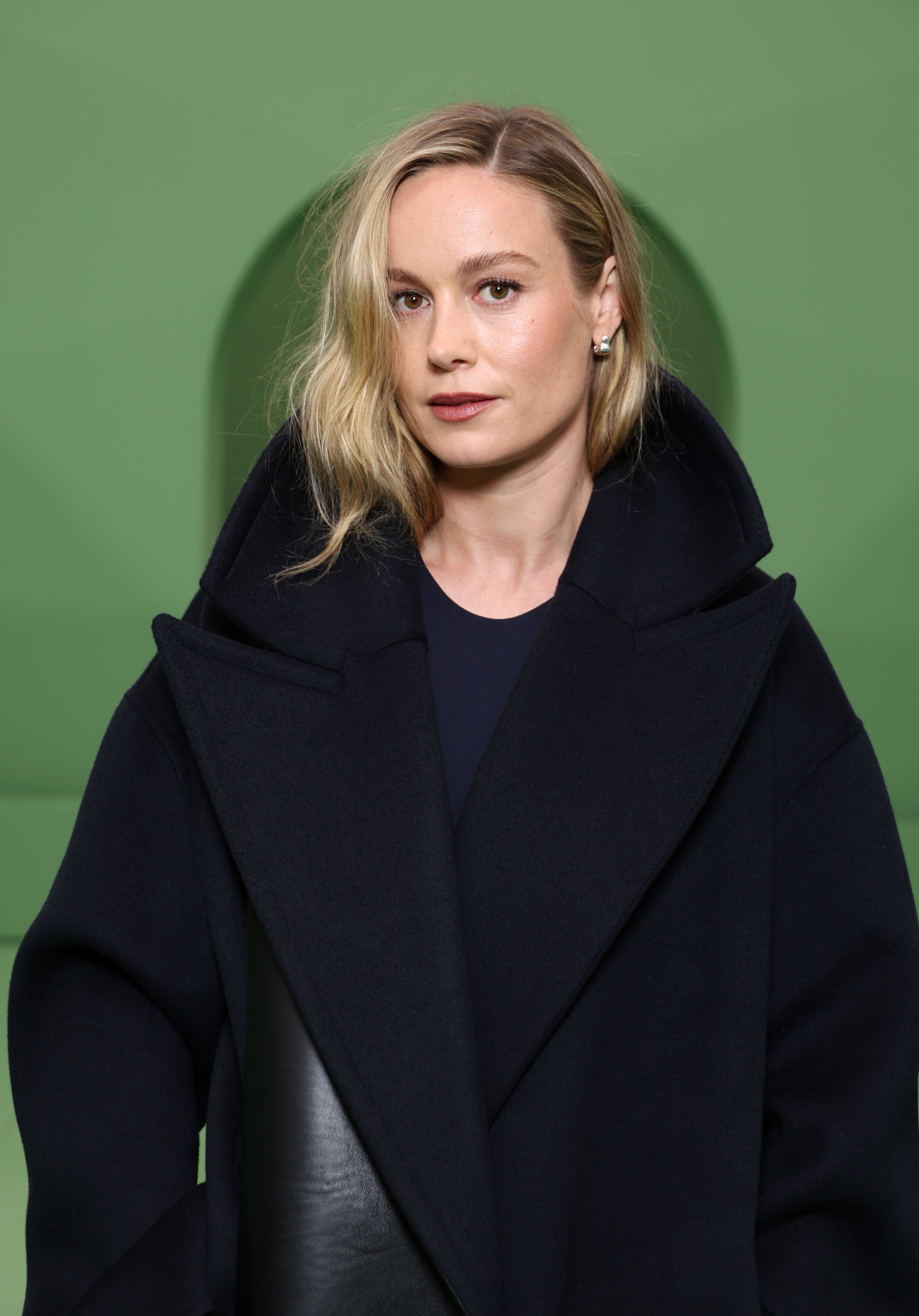 Brie Larson wearing a large black coat poses for a photo against a green background