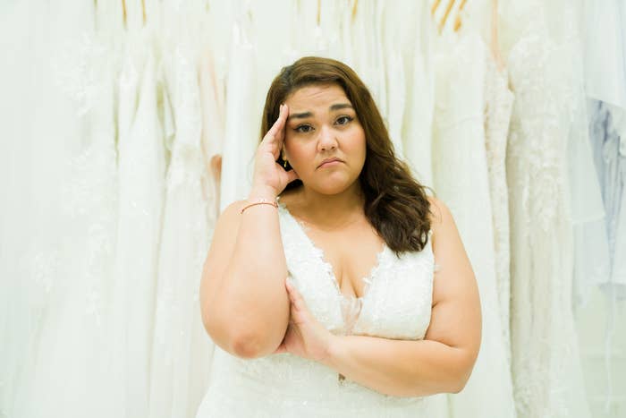 A person in a sleeveless, lace wedding dress stands among hanging bridal gowns, looking pensive with one hand on their face and the other arm crossed