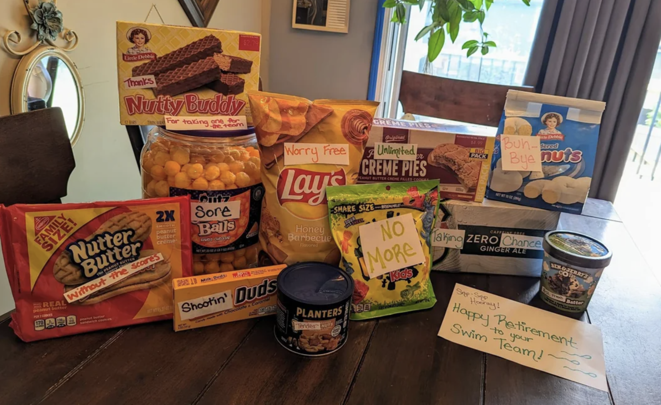 A table is covered with various snacks, including Nutty Buddy, Lay's chips, Little Debbie Creme Pies, and more. Two cards say, "Happy Retirement to your swim team!" and "No more chips!"