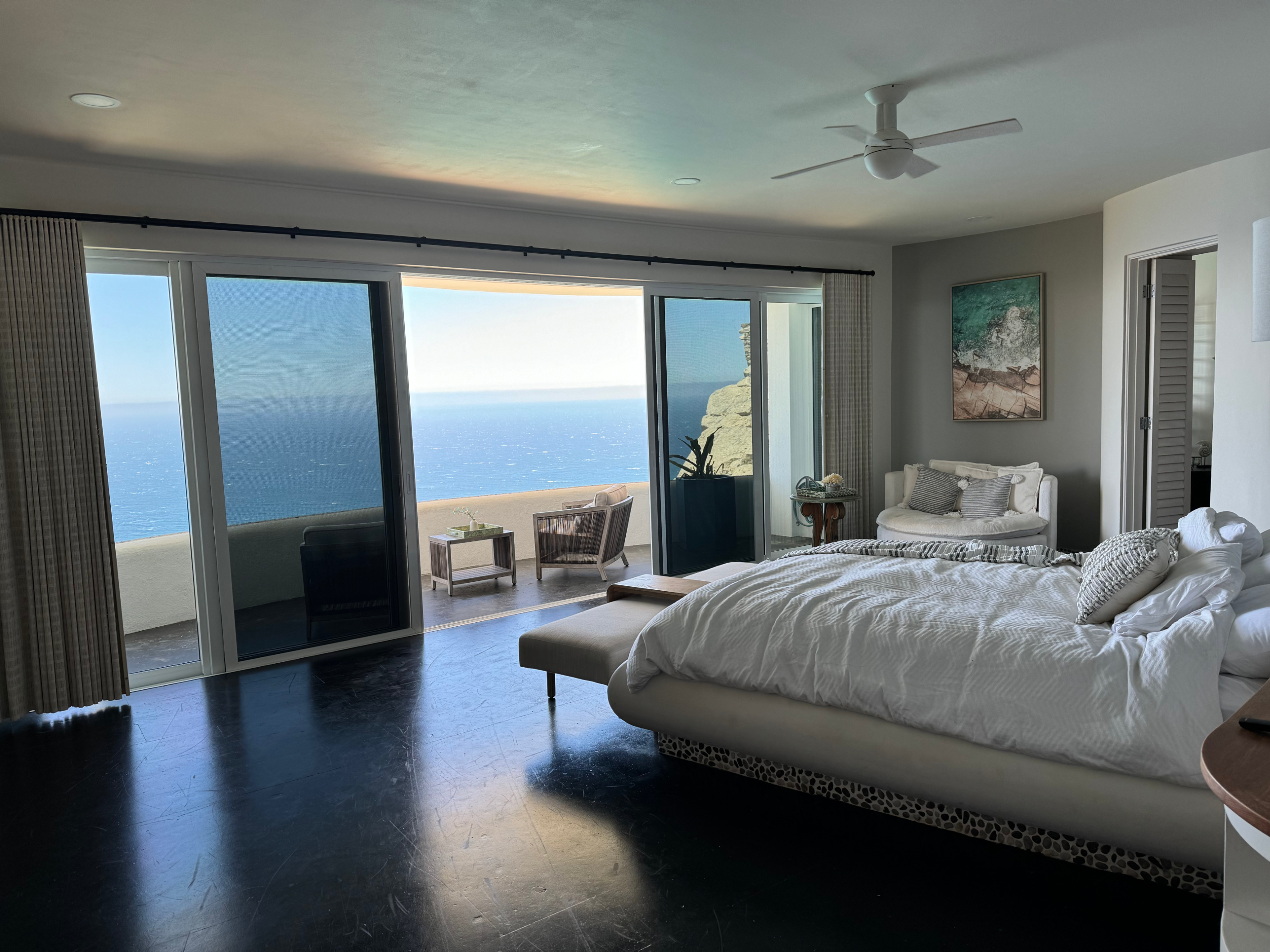 Spacious bedroom with a large bed, seating area, and floor-to-ceiling windows opening to a balcony with ocean views