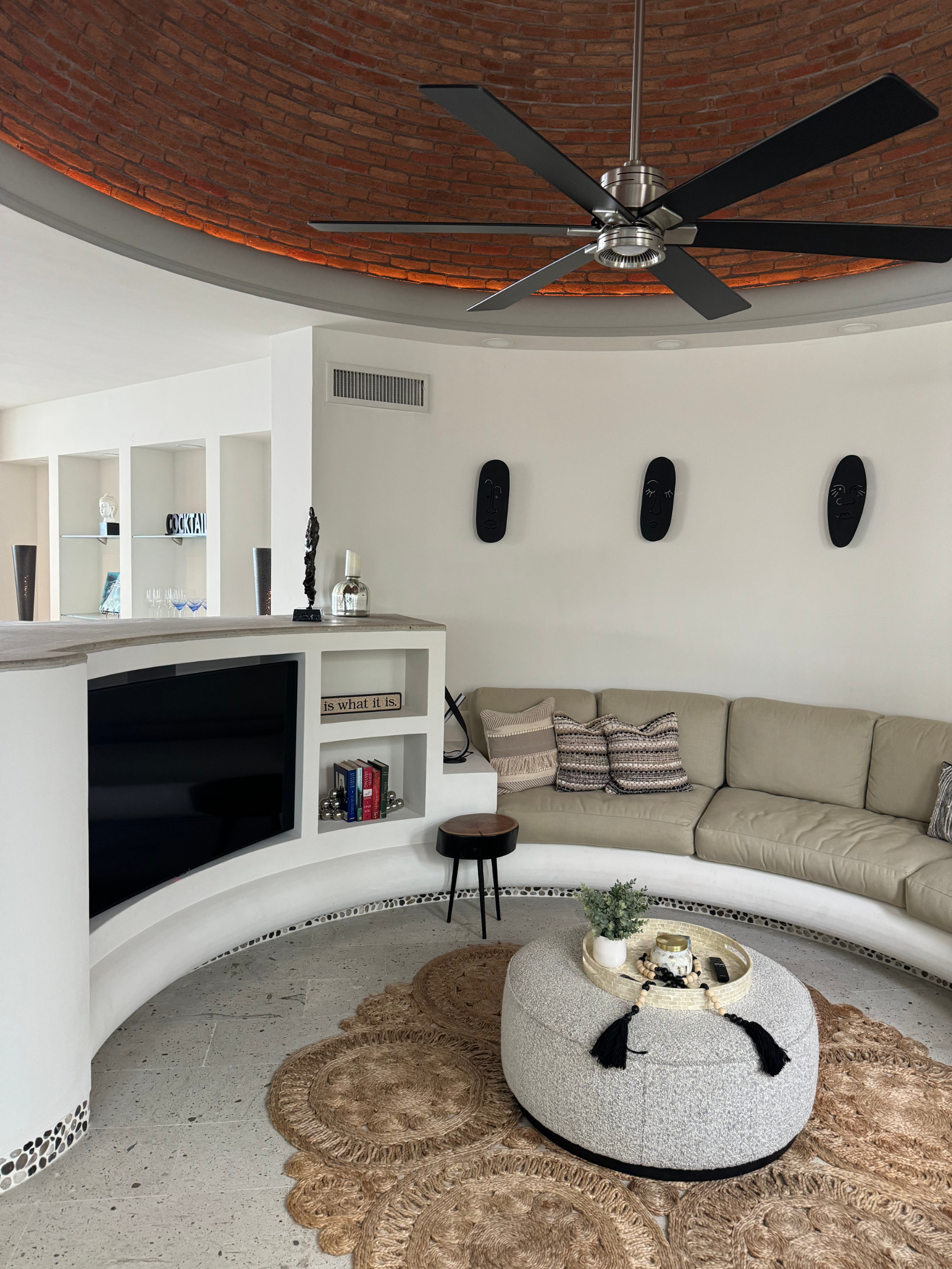 Modern living room with a curved sofa, round coffee table, built-in TV, and a ceiling fan. The room features a brick dome ceiling and decorative pillows
