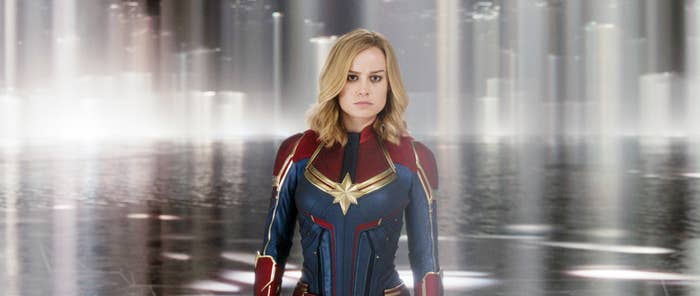 Brie Larson as Captain Marvel, wearing a superhero suit with prominent gold star emblem, in a futuristic, glowing background setting