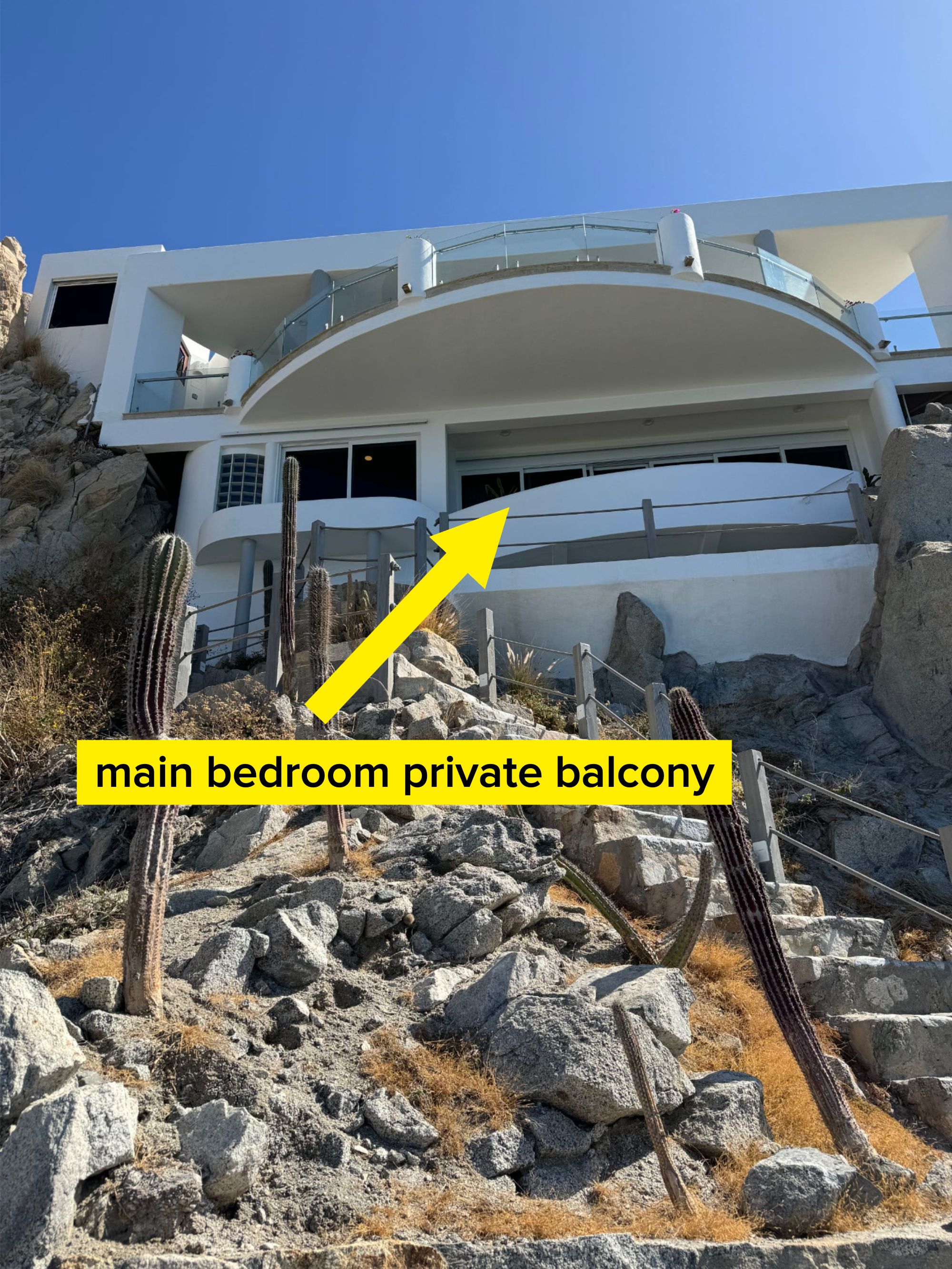 Modern white building with balconies built on a rocky hillside with cacti in the foreground, under a clear sky