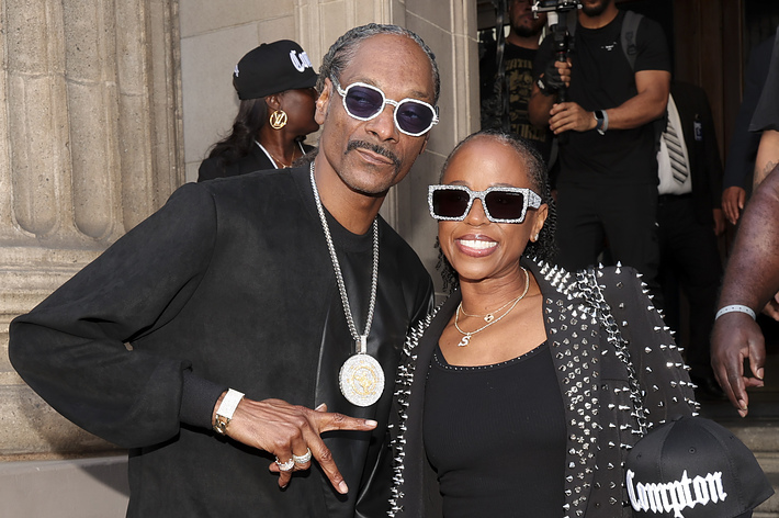 Snoop Dogg and Sheryl Lee Ralph pose together holding hands. Snoop Dogg wears a black outfit with a large medallion, while Ralph wears sunglasses and a studded jacket