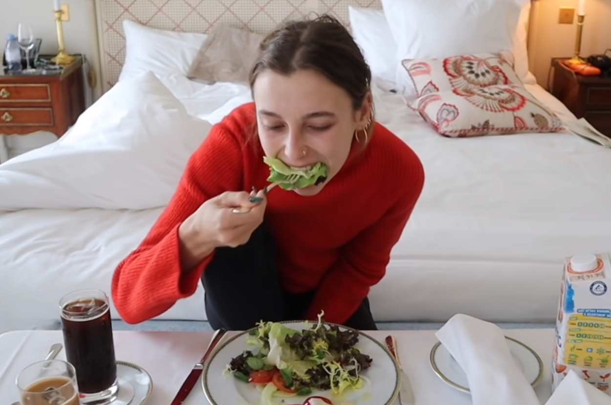 Emma Chamberlain, sitting on a bed in a hotel room, eating a salad with a fork