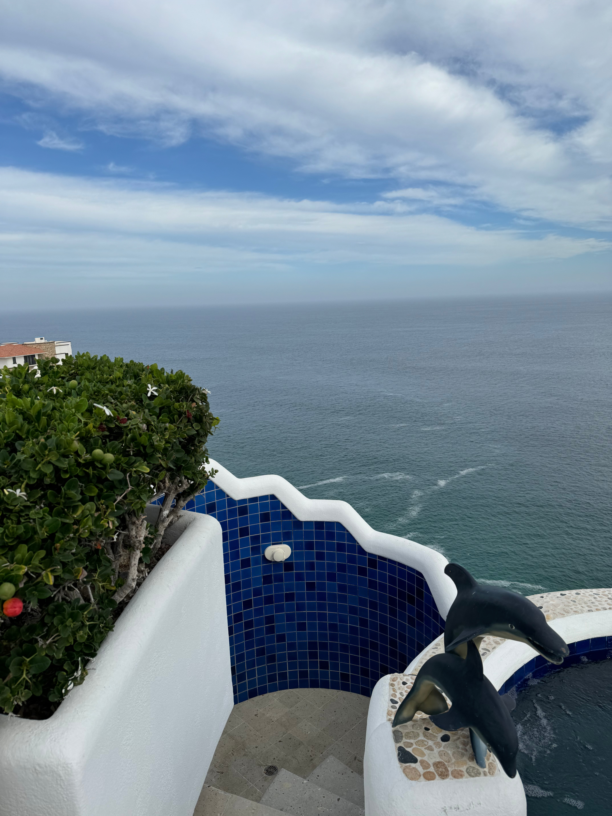 Ocean view from a white and blue-tiled balcony featuring a decorative dolphin statue and a green bush. The balcony overlooks a vast expanse of water under a cloudy sky