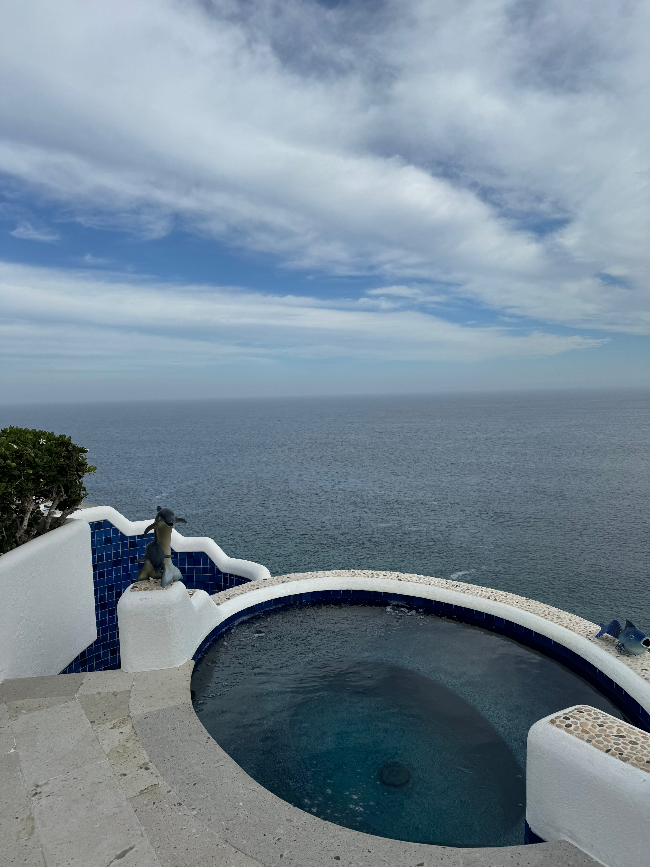 Infinity pool overlooking a calm ocean, with blue tiles and a statue of a dolphin nearby. Sky is overcast, adding a tranquil atmosphere to the scene