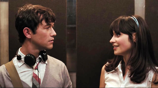 Joseph Gordon-Levitt and Zooey Deschanel in an elevator scene. Joseph is wearing a shirt and tie, with headphones around his neck. Zooey wears a headband and sleeveless top
