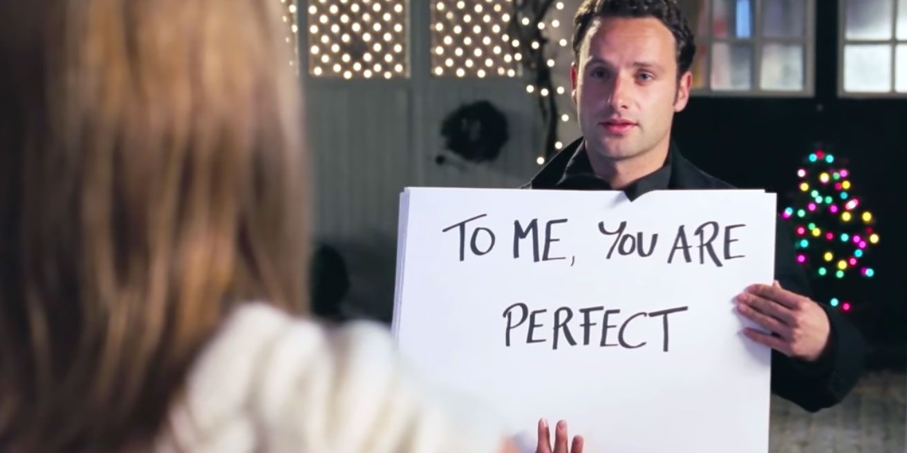 Andrew Lincoln holds a sign reading "TO ME, YOU ARE PERFECT" to a woman with her back to the camera, in a scene with holiday lights