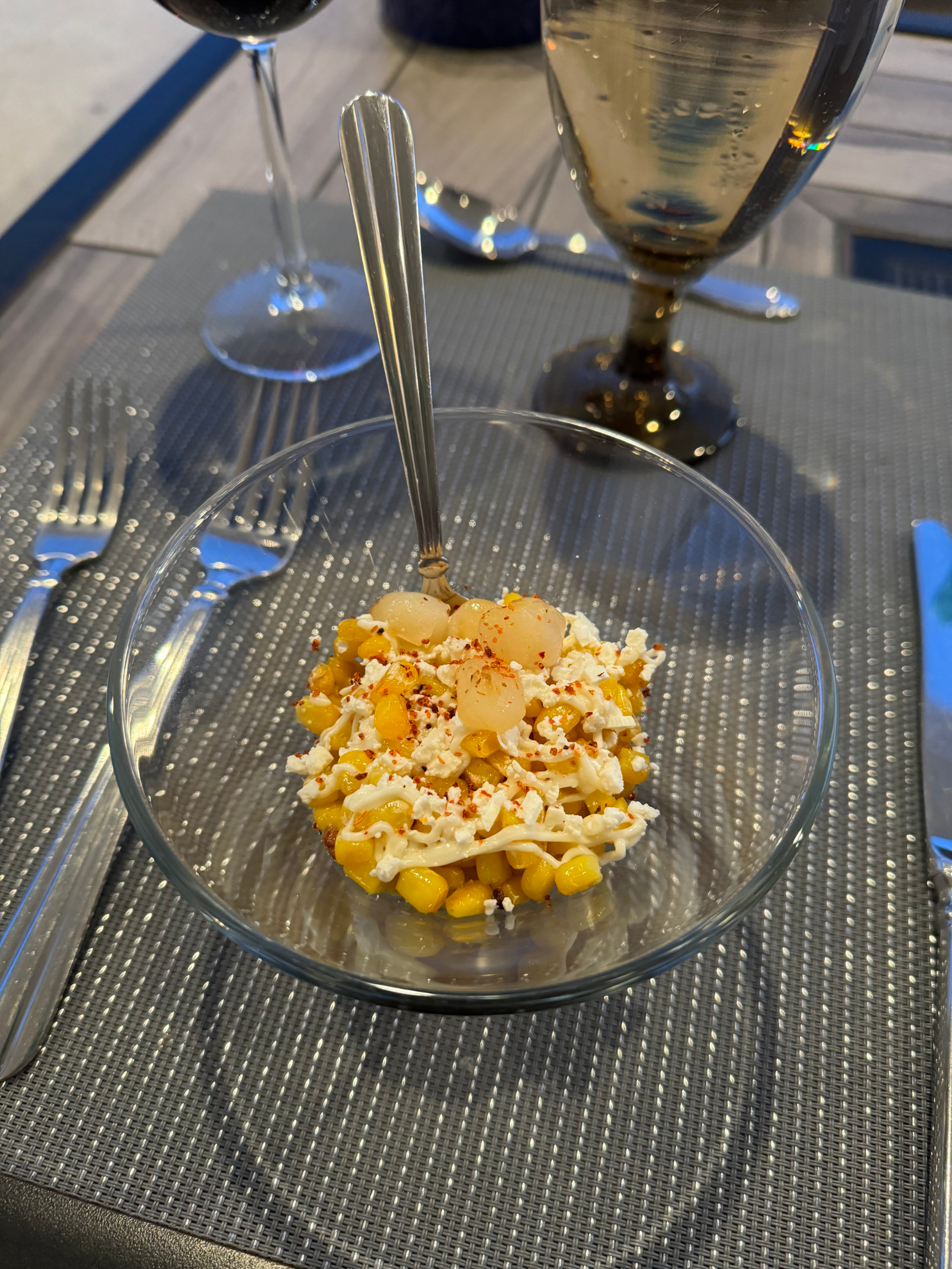 A bowl of corn salad topped with onions and spices, placed on a set dining table with a knife, fork, wine glass, and water glass