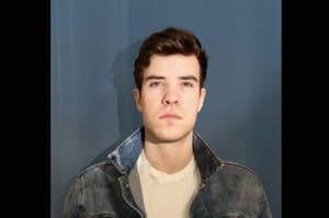 A young man poses for a photo against a plain background, wearing a denim jacket over a white shirt, with a neutral expression on his face. Name unknown