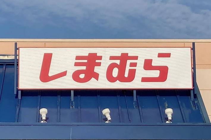 The image shows the exterior of a store with a sign in Japanese characters above the entrance, reading &ldquo;しまむら.&rdquo; There is an accessible parking sign visible