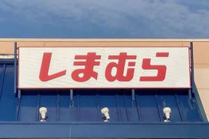 The image shows the exterior of a store with a sign in Japanese characters above the entrance, reading "しまむら." There is an accessible parking sign visible