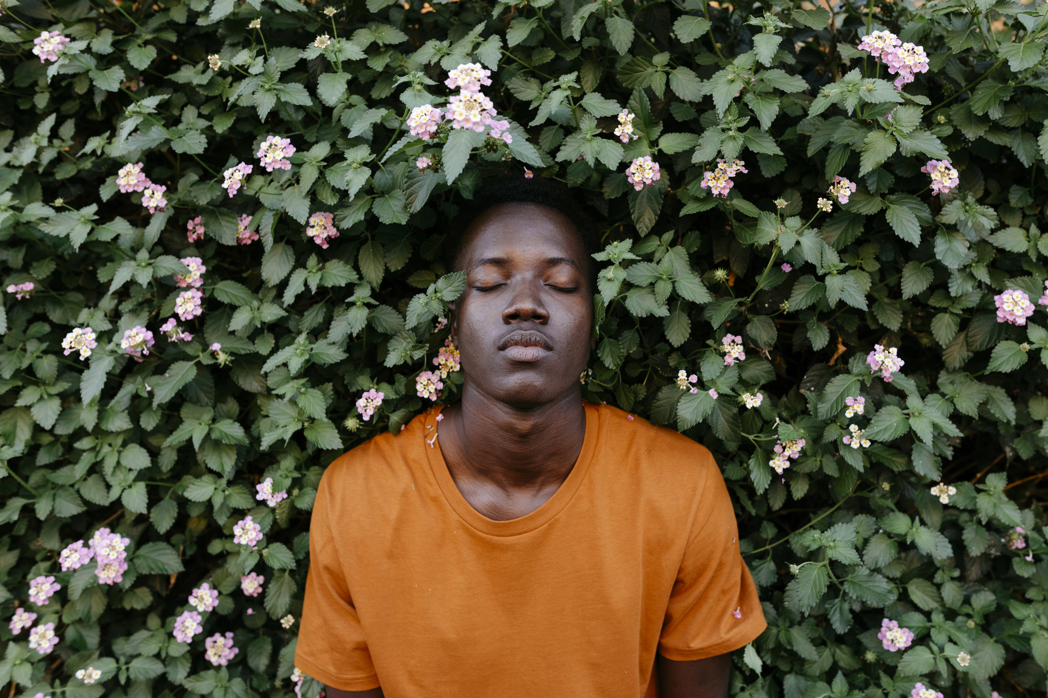 Person in an orange shirt stands against greenery with pink flowers, eyes closed, appearing tranquil