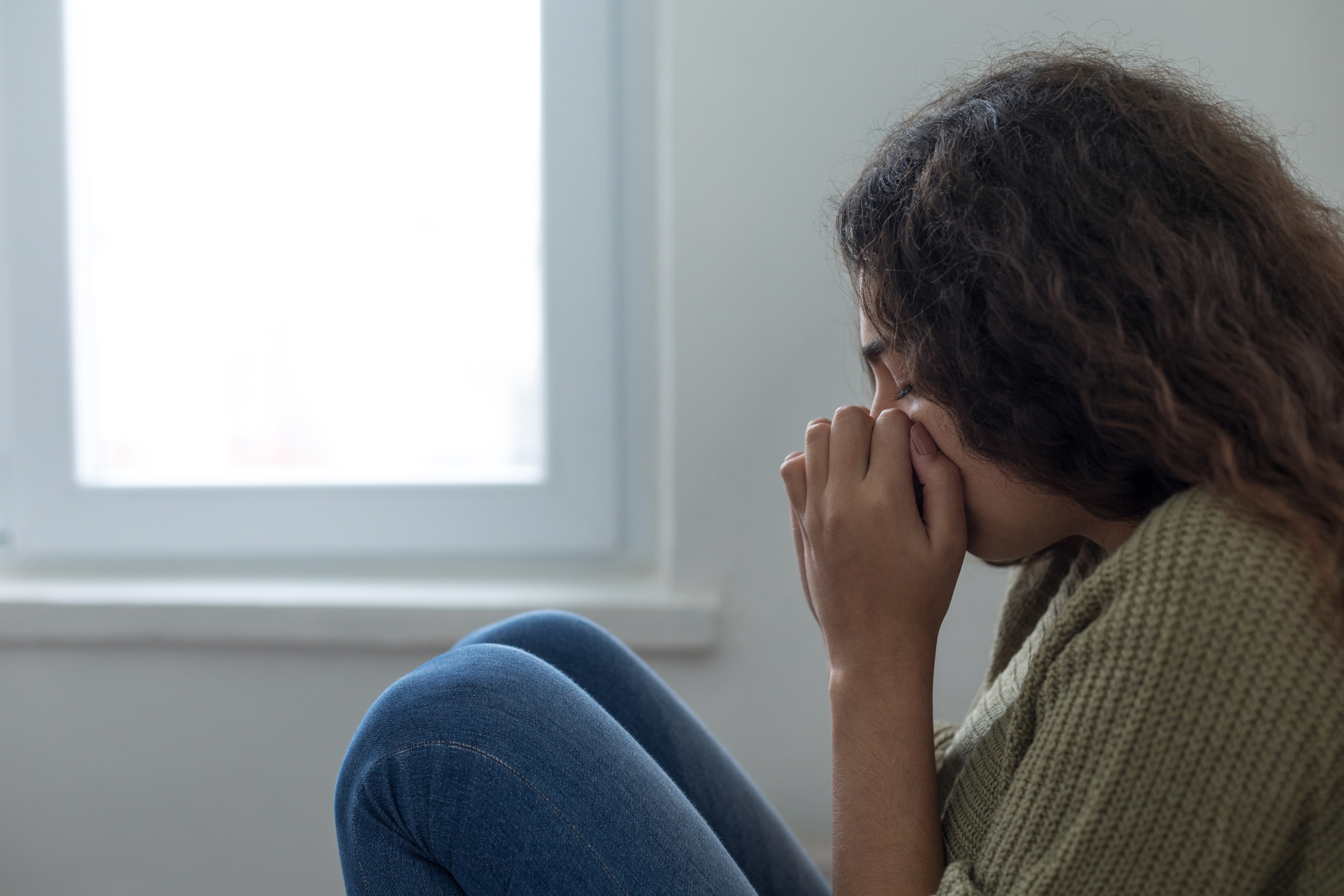 A woman sits by a window, appearing upset with her hands covering her face