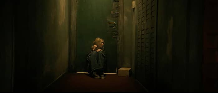 Young woman crouches in a dimly lit hallway, leaning against a green door. A small cardboard package is placed on the floor beside her