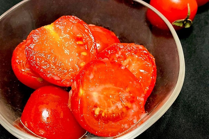 透明なボウルにいくつかのスライスされたトマトと丸ごとのトマトが盛られている。背景には丸ごとのトマトが3つ見える。