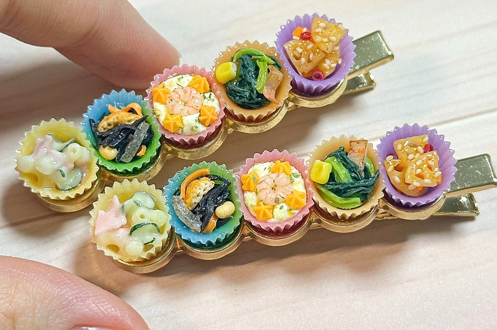 手作りのミニチュア食品がデザインされたヘアクリップを持っている手の画像。