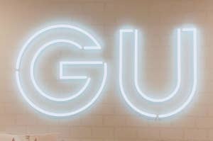 Large glowing "GU" sign on a wall with several white shopping bags below it, also branded with the "GU" logo