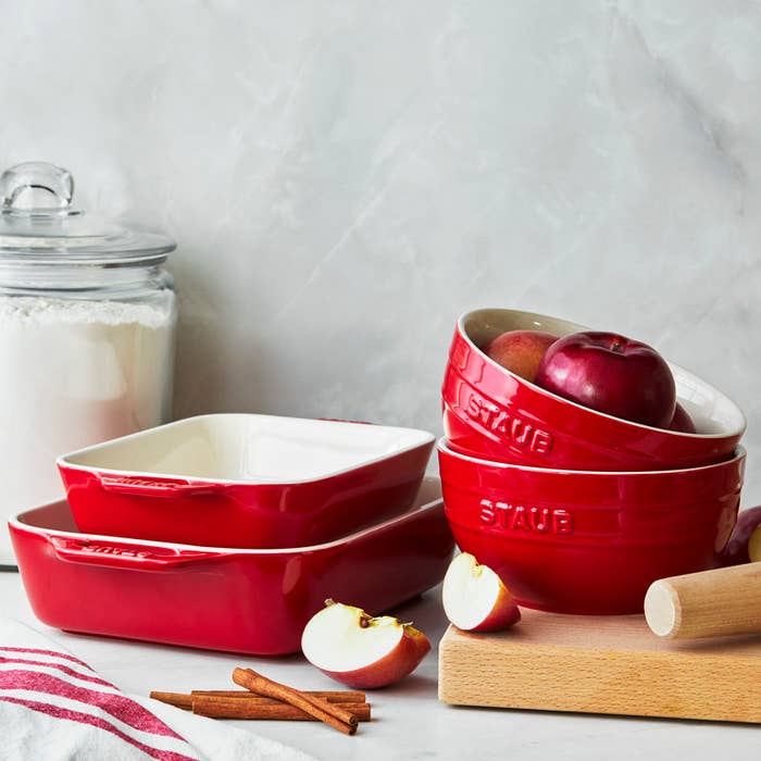 Stacked red ceramic cookware on a counter with apples and a rolling pin nearby