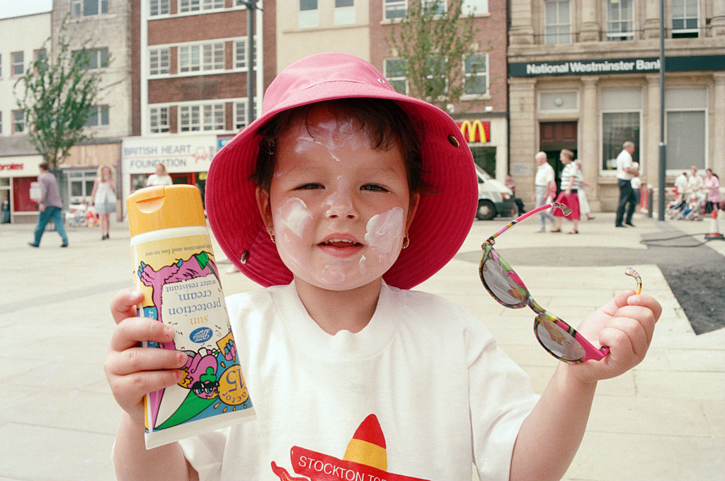 Child with sunscreen on face holding a juice box and sunglasses, wearing a sun hat and T-shirt at a sunny outdoor event