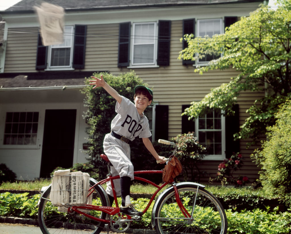 A paperboy in vintage attire throws a newspaper while riding a bike