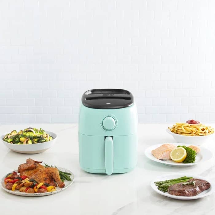 Compact air fryer on a kitchen counter with assorted dishes including vegetables, fries, fish, and steak