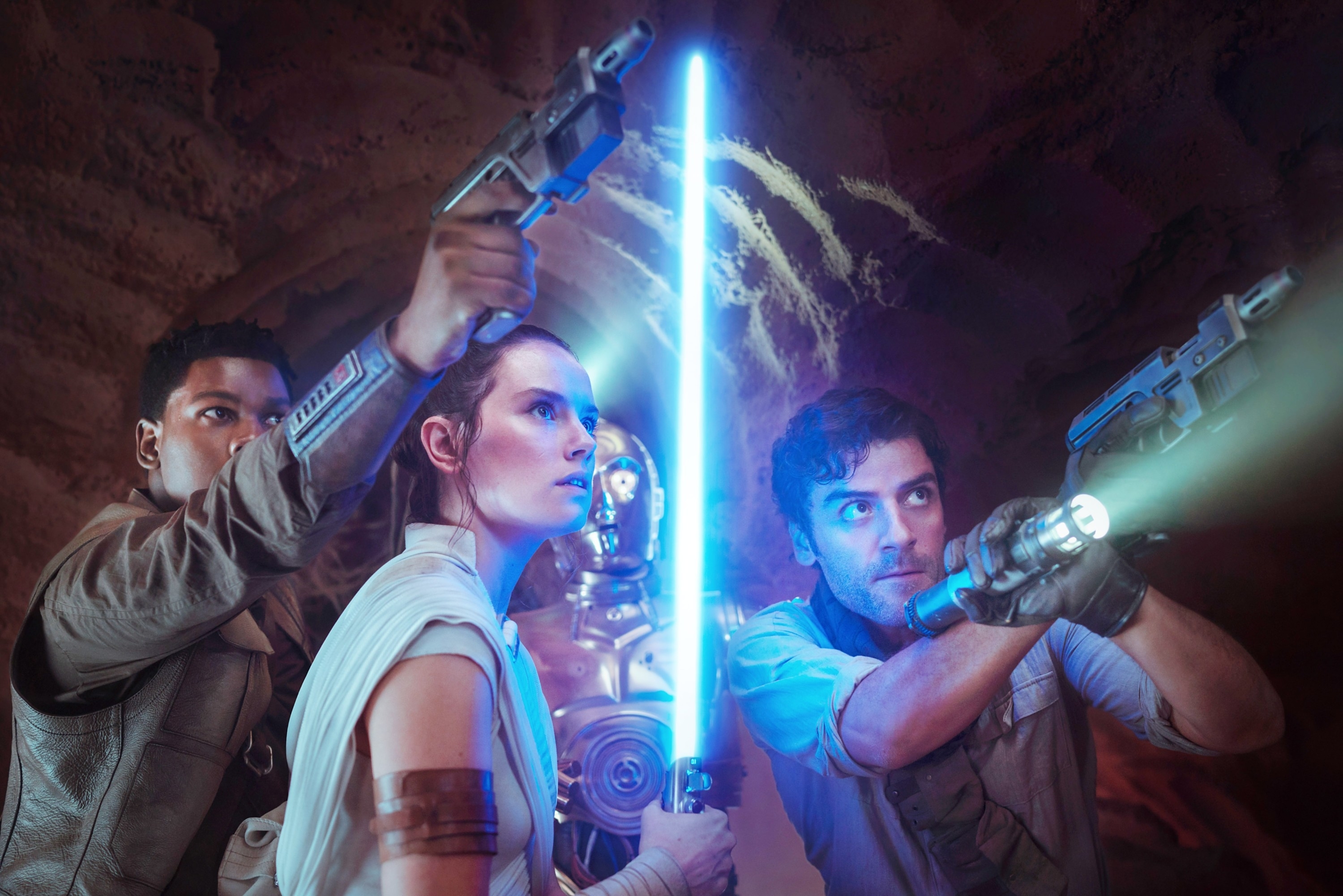 John Boyega, Daisy Ridley, and Oscar Isaac as their Star Wars characters ready for action. Ridley holds a lightsaber