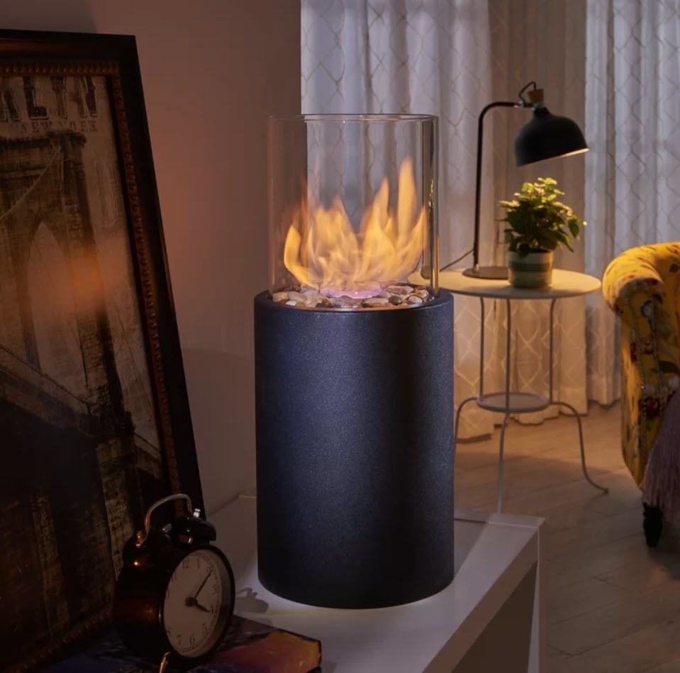 Indoor portable fireplace with visible flames, set on a cylindrical base next to a window