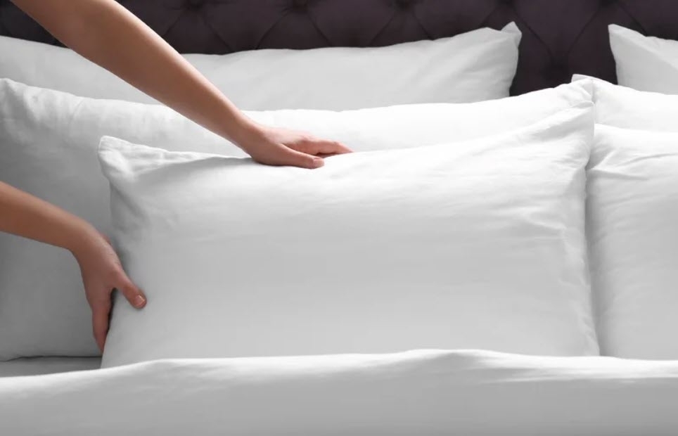 A person&#x27;s hands smoothing a white pillow on a neatly made bed