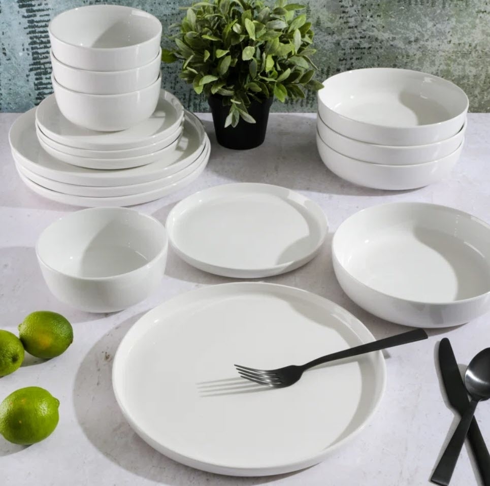 Various white dinnerware items and utensils are neatly arranged on a table, accompanied by a plant and limes