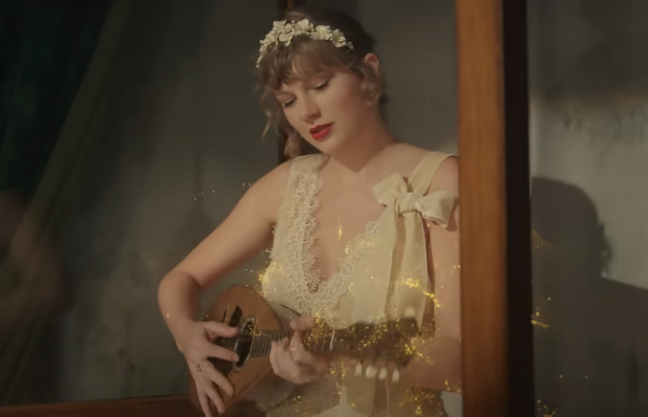 Taylor Swift plays a guitar, wearing a vintage lace dress with a floral headpiece