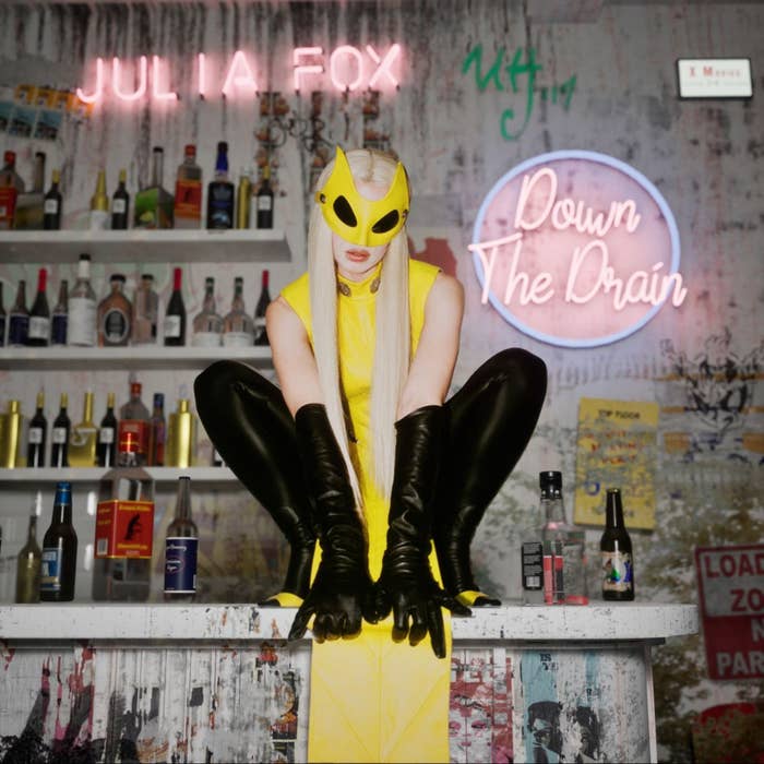 Person in yellow attire and mask crouches on counter with neon signs and bottles in background