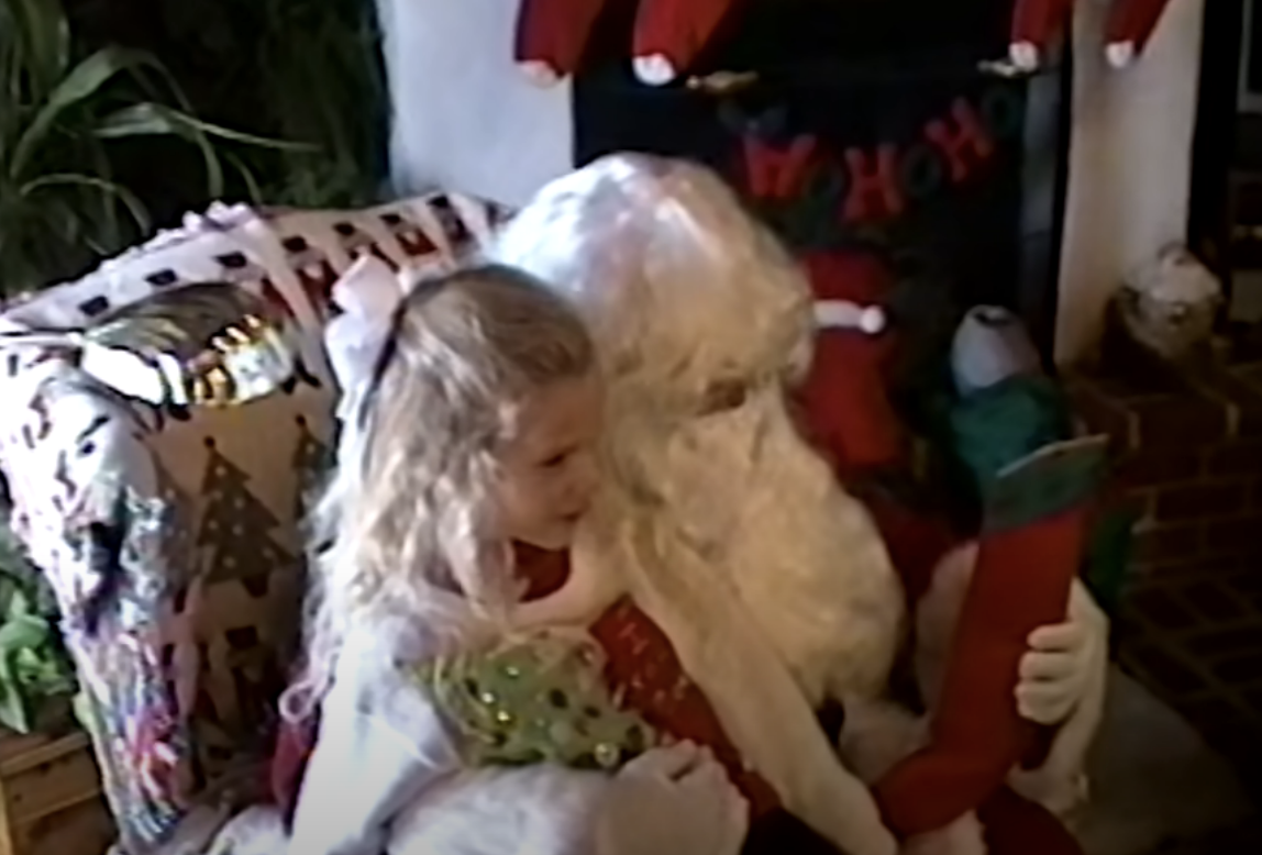 A child sits on Santa&#x27;s lap by a fireplace with stockings, holding a stocking and conversing