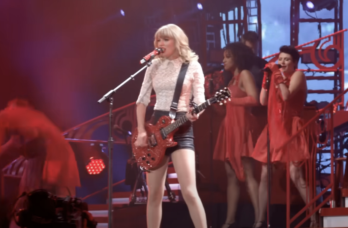 Taylor Swift performs onstage with a guitar, flanked by backup singers