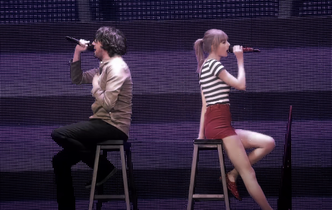 Two performers on stage with microphones, seated on stools against a lit backdrop