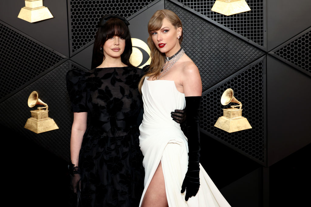 Two women at an awards event, one in a black dress with sheer elements, the other in a white gown with a high slit, standing near award statues