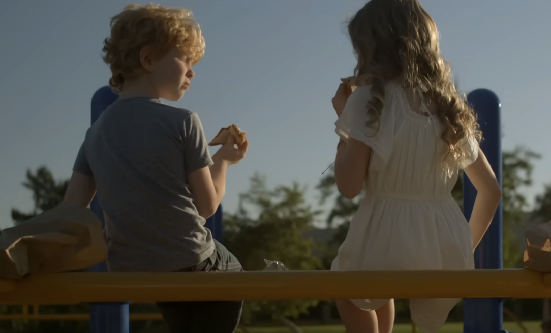 Two children eating sandwiches on a park bench, facing away from the camera