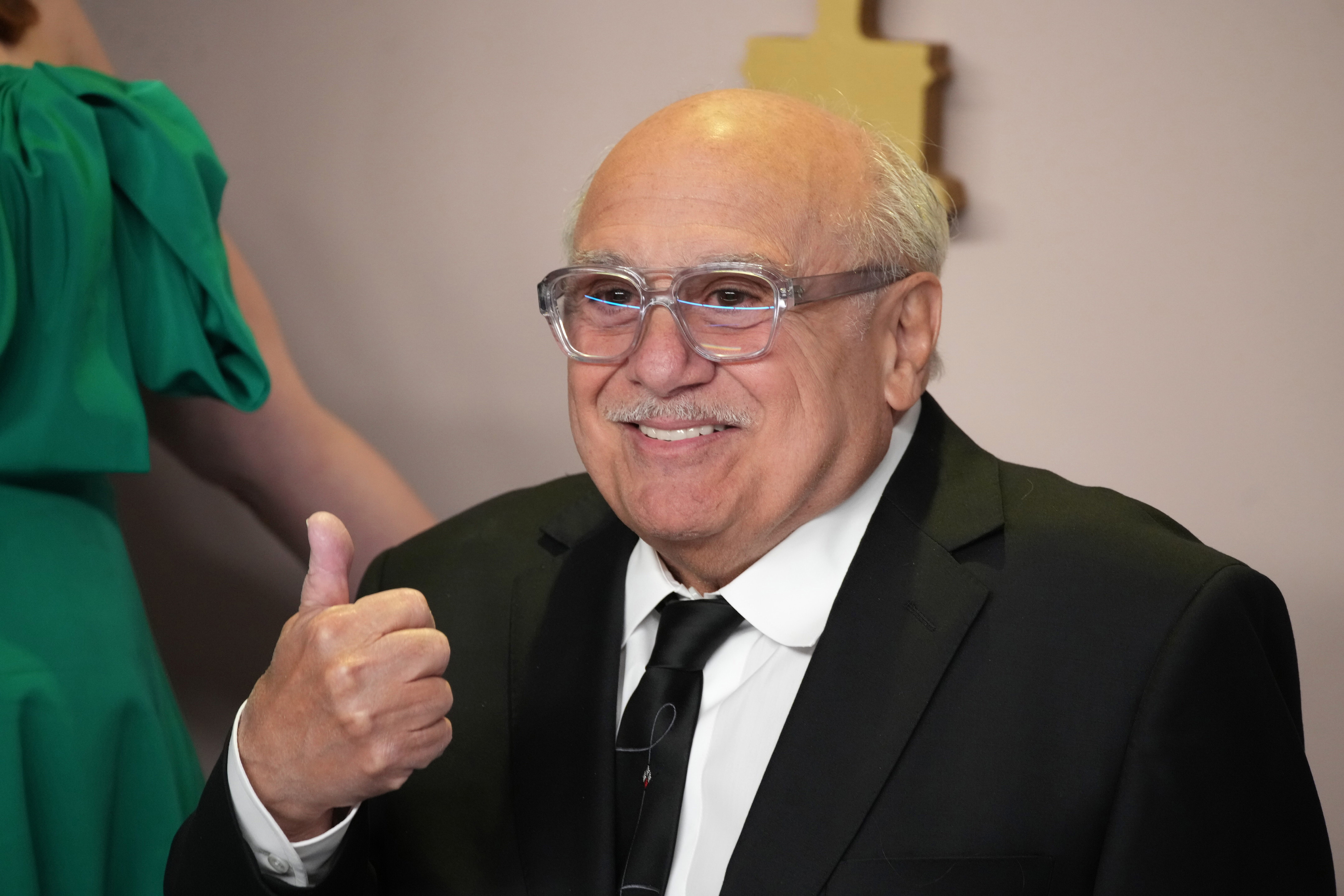 Danny DeVito in a black suit gives a thumbs up at an event